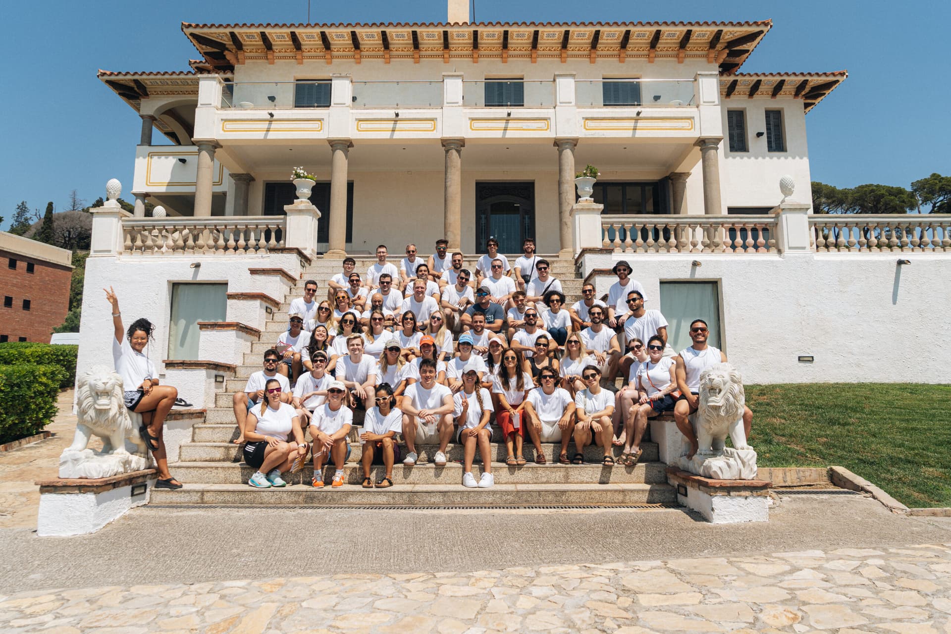 A large group of approximately 40-50 people, mostly wearing white t-shirts, posing for a group photo on the wide stone steps of an elegant, multi-story Mediterranean-style villa with ornate balconies, columns, and decorative railings under a clear blue sky. The group is arranged in several rows, smiling and looking at the camera, with two large white stone lion statues flanking the staircase on either side, set against a backdrop of green lawn and trees, conveying a sense of team camaraderie and a successful corporate offsite event.