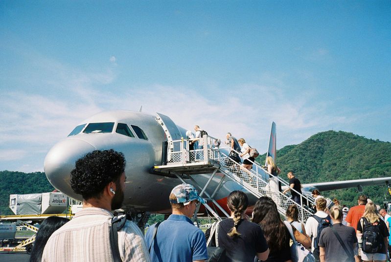 A wide panoramic image showing a group of travelers boarding a large commercial airplane via a mobile staircase under a clear blue sky, with lush green hills in the background, suggesting a corporate retreat or group travel scenario.