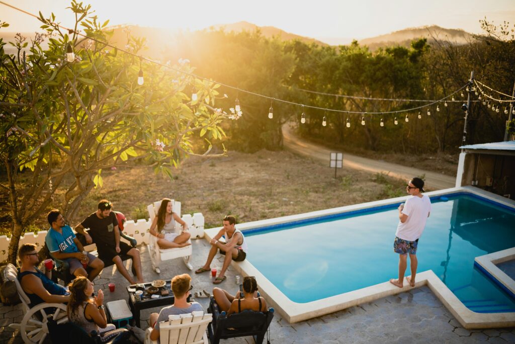 A warm, golden-hour scene of a group of people enjoying a relaxed outdoor gathering beside a rectangular swimming pool, with string lights hanging overhead, lush greenery, and distant hills in the background, capturing the essence of a company summer event or corporate party.