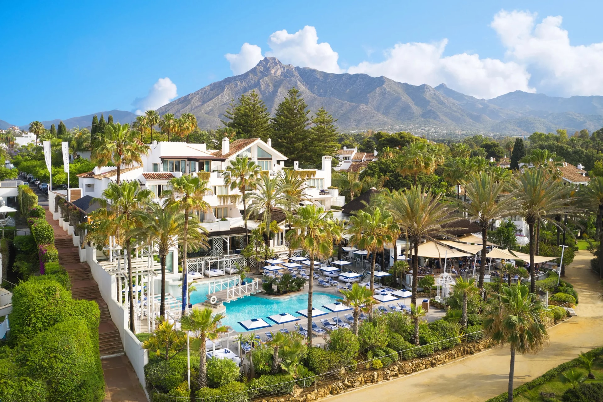 A wide panoramic image showcasing a luxurious resort in Marbella, Spain, featuring white Mediterranean-style buildings with terracotta roofs, surrounded by lush greenery and tall palm trees. A vibrant blue swimming pool with numerous sun loungers and blue-and-white umbrellas is centrally located, while a majestic mountain range with rugged peaks dominates the background under a clear blue sky with scattered white clouds. This scenic view highlights the resort's blend of natural beauty and upscale amenities, ideal for leisure and corporate retreats.
