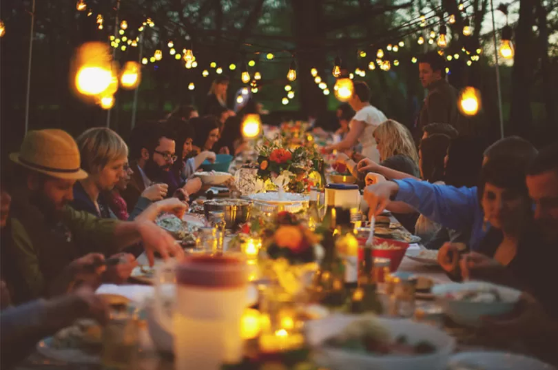 A warm, festive outdoor dinner party scene at dusk, featuring a long table laden with food, drinks, and floral centerpieces, surrounded by diverse guests enjoying a meal together under strings of glowing fairy lights and lanterns, evoking a cozy, communal summer fiesta atmosphere.