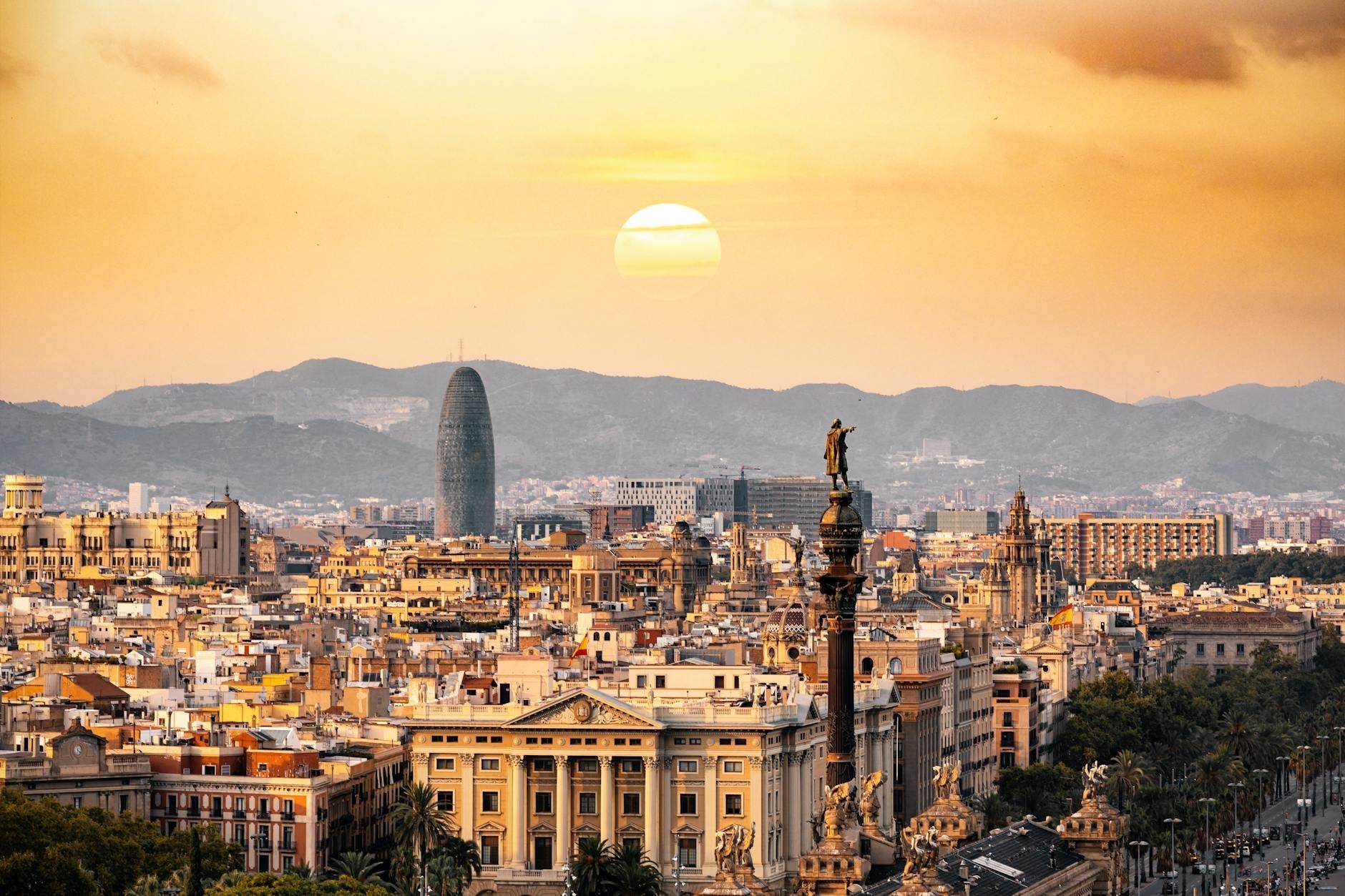 A panoramic sunset view of Barcelona’s skyline bathed in golden hour light, featuring the iconic Torre Glòries (formerly Torre Agbar) rising prominently in the midground, the Columbus Monument with its bronze statue pointing toward the sea in the foreground, and a dense tapestry of historic and modern buildings stretching across the cityscape; distant hills frame the horizon under a warm orange sky with a large, glowing sun partially obscured by haze, while palm-lined avenues and classical architecture add texture and depth to the urban vista.